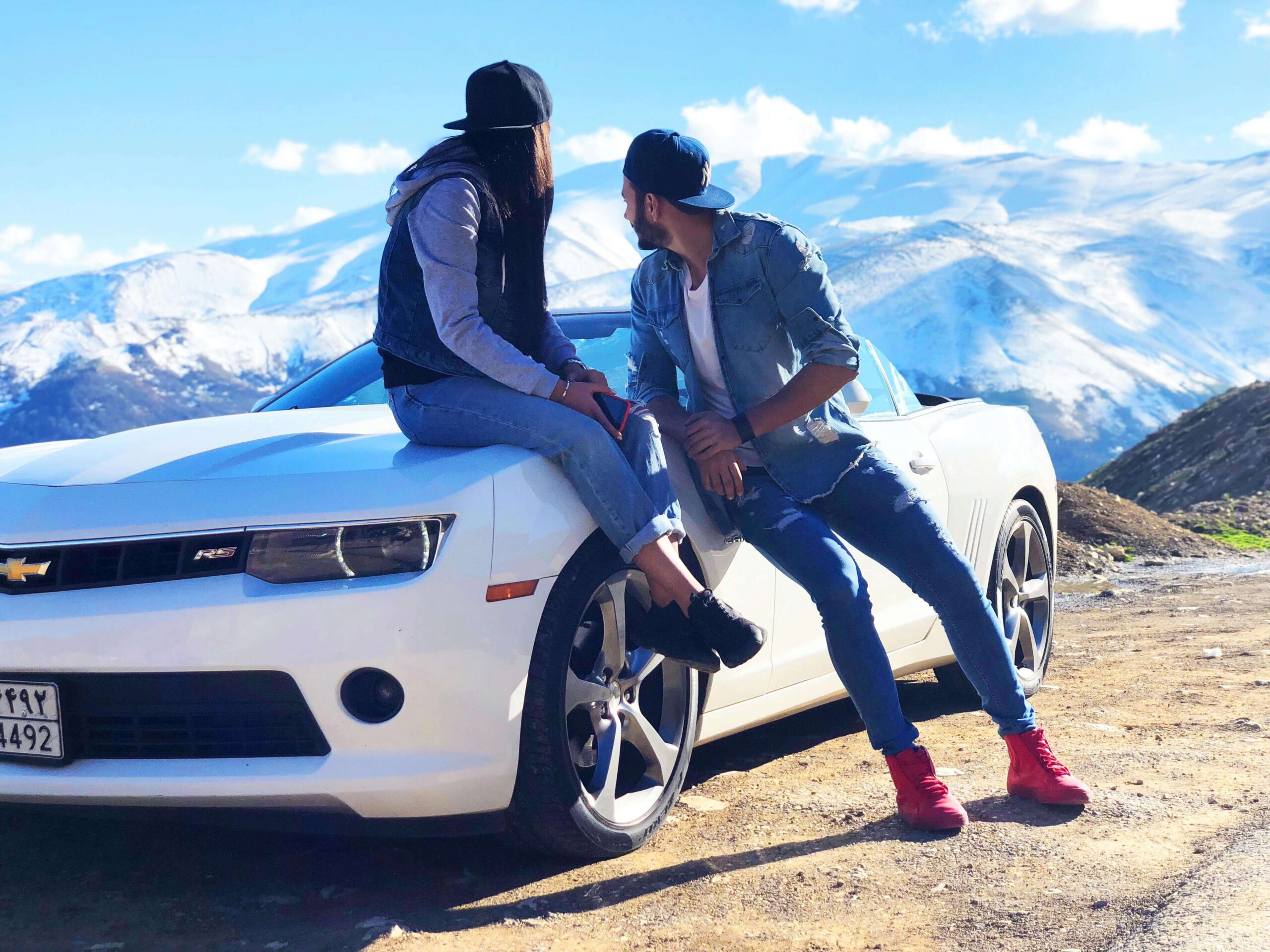 A couple enjoying a moment on the hood of their white convertible car with snow-capped mountains in the background
