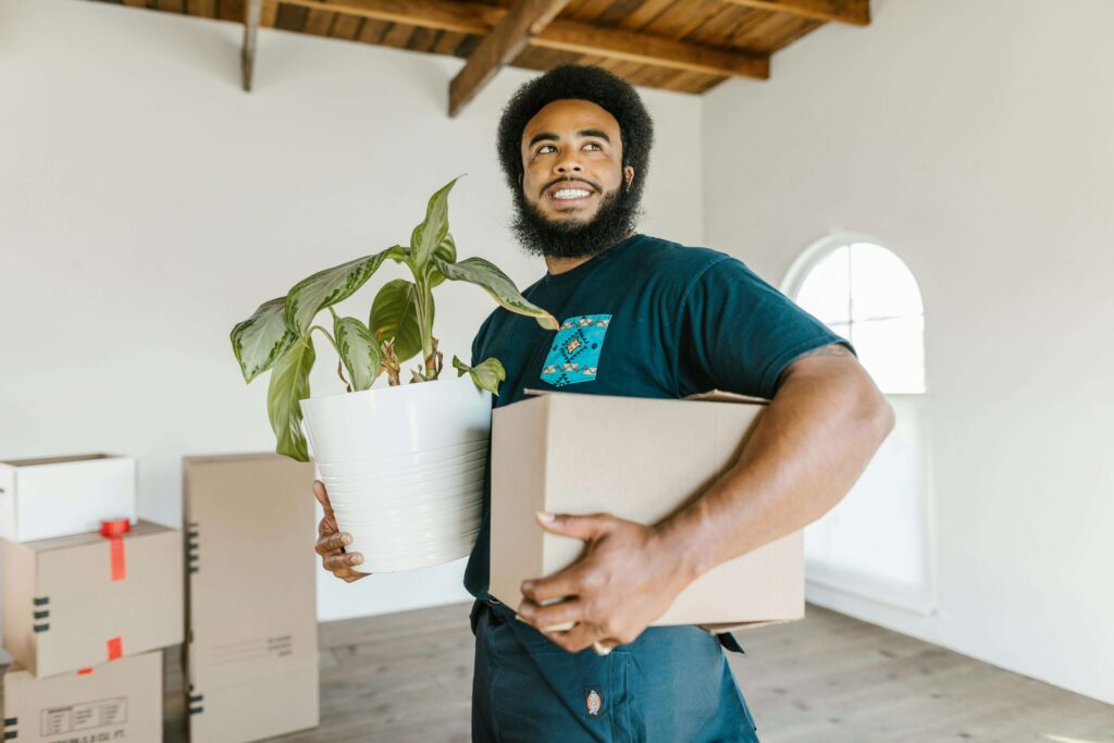 A mover with a friendly smile carrying a cardboard box and an office plant, ensuring a safe transfer