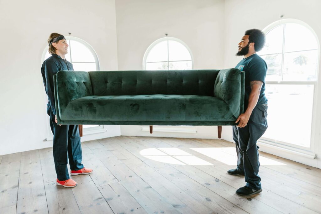 Two professional movers carefully carrying a large, dark green velvet sofa in a bright, empty room