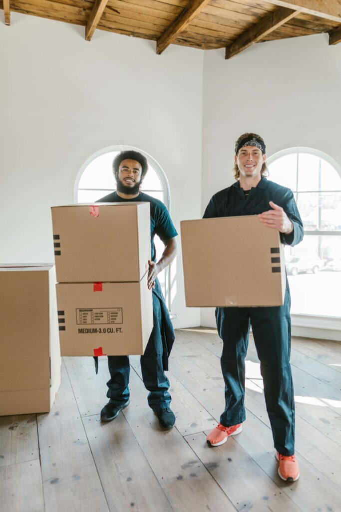 Two smiling movers carrying cardboard boxes in a well-lit office space.
