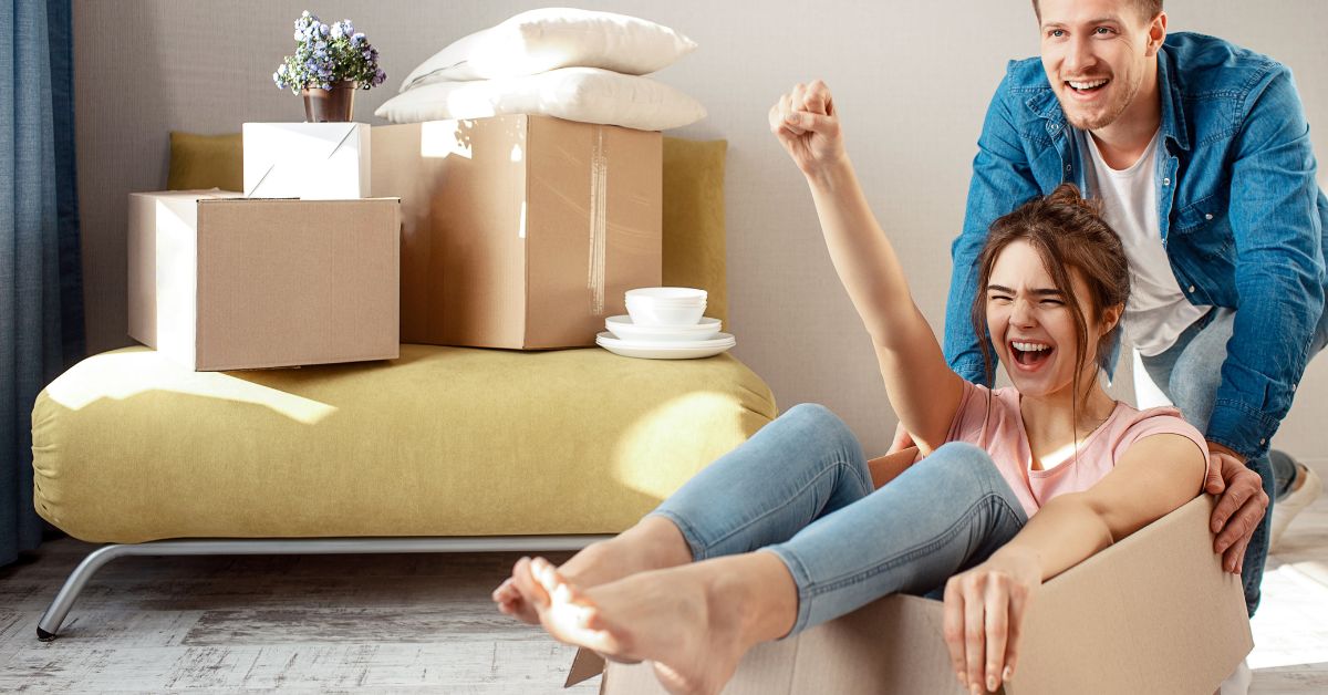 A happy couple enjoying their move; the man pushes the woman in a cardboard box, surrounded by packed items.